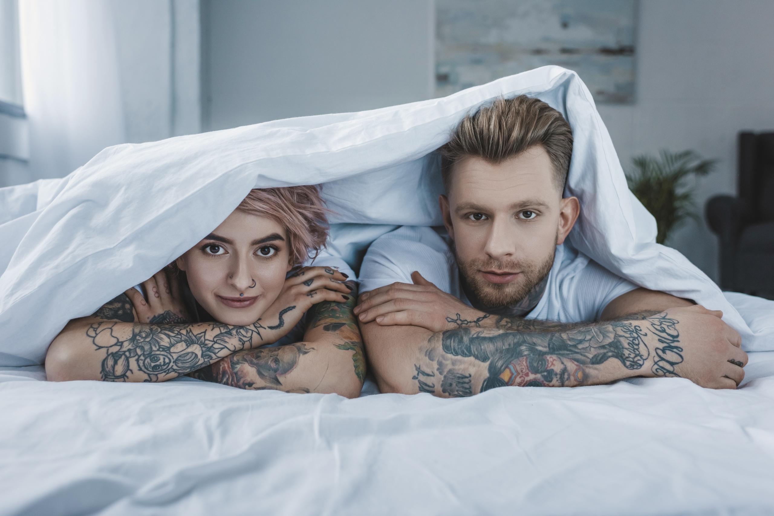 Young,Tattooed,Couple,Lying,In,Bed,Under,White,Blanket